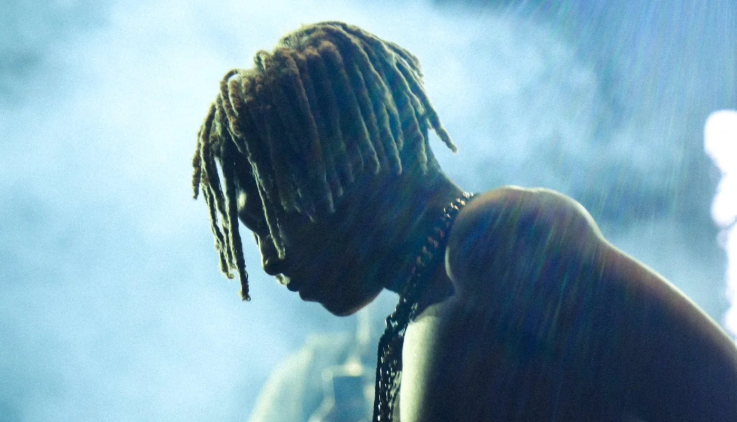 Primo piano di profilo di XXXTENTACION con la testa china e una catena al collo, tra fumo e luci blu