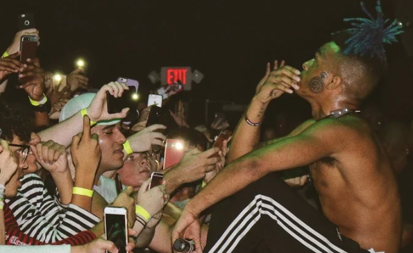 XXXTENTACION seduto sul bordo del palco con la mano al viso, circondato da fan che lo riprendono con i telefoni