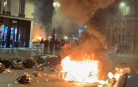 Cassonetti e rifiuti in fiamme in una strada di città di notte, con alcune persone sullo sfondo: immagine di una rivolta di piazza