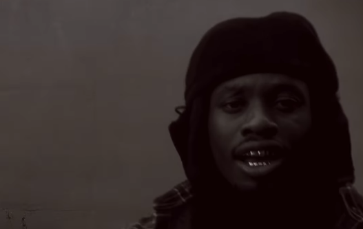 Fotogramma di Denzel Curry con cappuccio e denti dorati, dal mondo visivo di ii